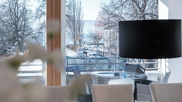 A modern hotel room with a large window offering a view of a cityscape, including a bridge and boats. - Exclusive new apartments with a romantic location at the small sailing port (Überlingen)