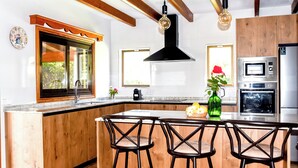 A modern kitchen with wooden cabinetry, a large island with bar stools, a built-in oven, and a window with a view of greenery. - Villa Can Lluc(Private villa with big barbecue) (Puerto de Pollença)