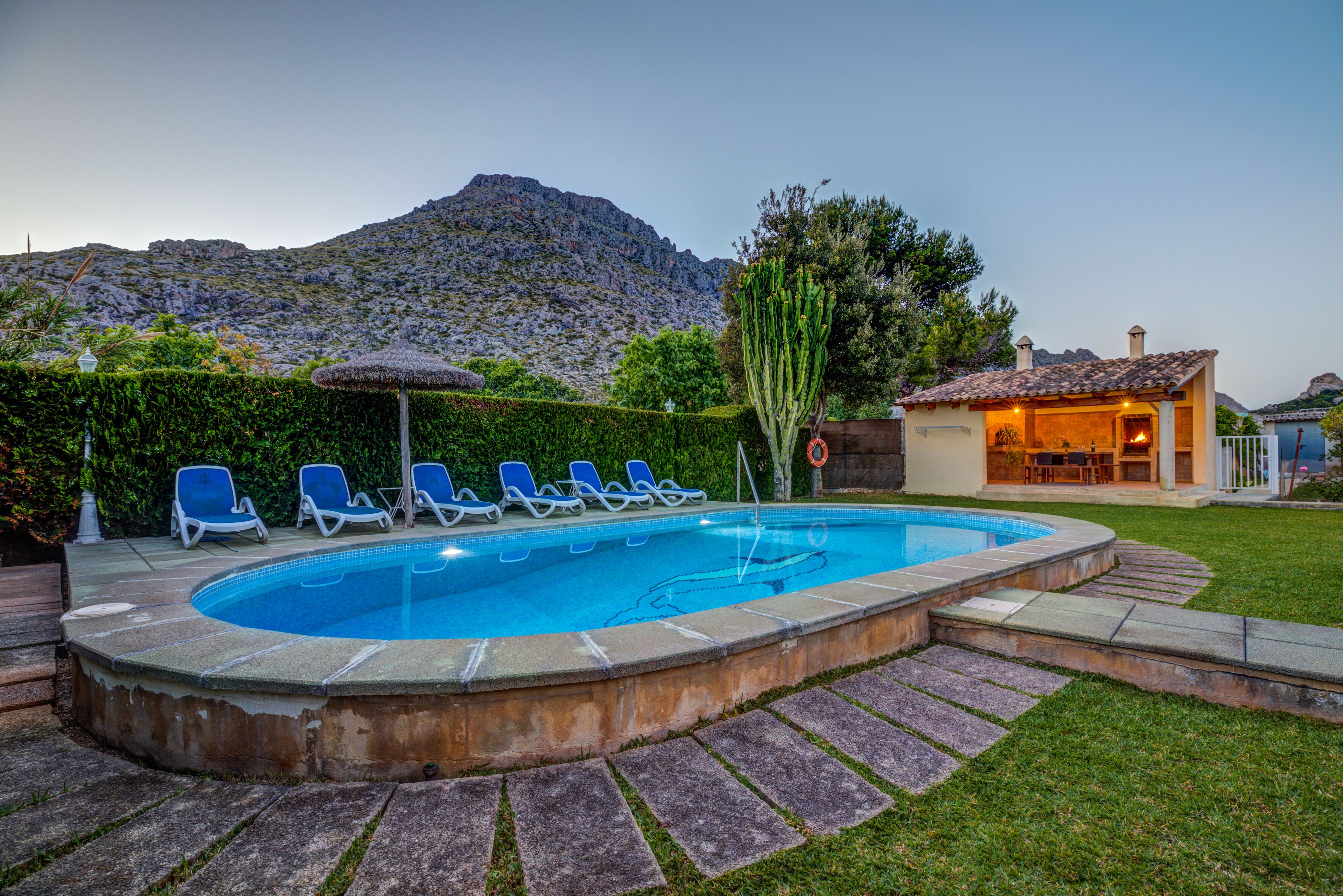 A swimming pool with lounge chairs, a small building with a tiled roof, and a mountainous backdrop.
