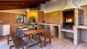 A rustic kitchen with a stone backsplash, wooden beams, a built-in grill, and a dining area with benches. - Villa Can Lluc(Private villa with big barbecue) (Puerto de Pollença)
