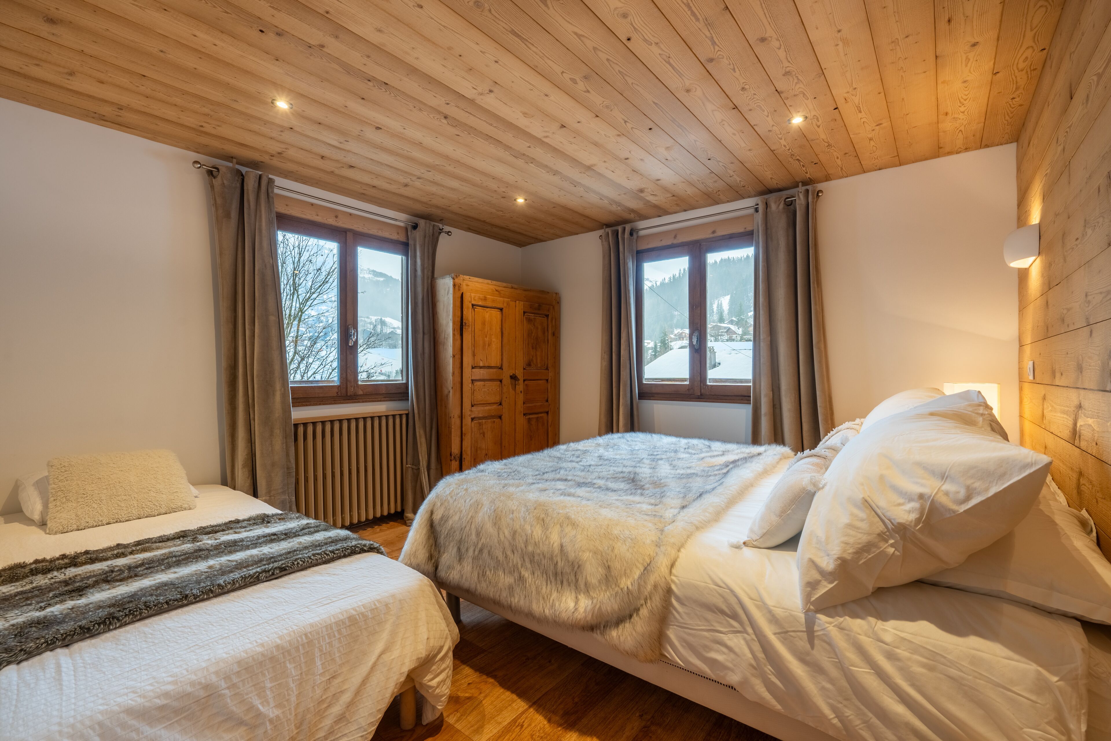 Een slaapkamer met twee bedden, een houten plafond, grote ramen en een houten garderobe.