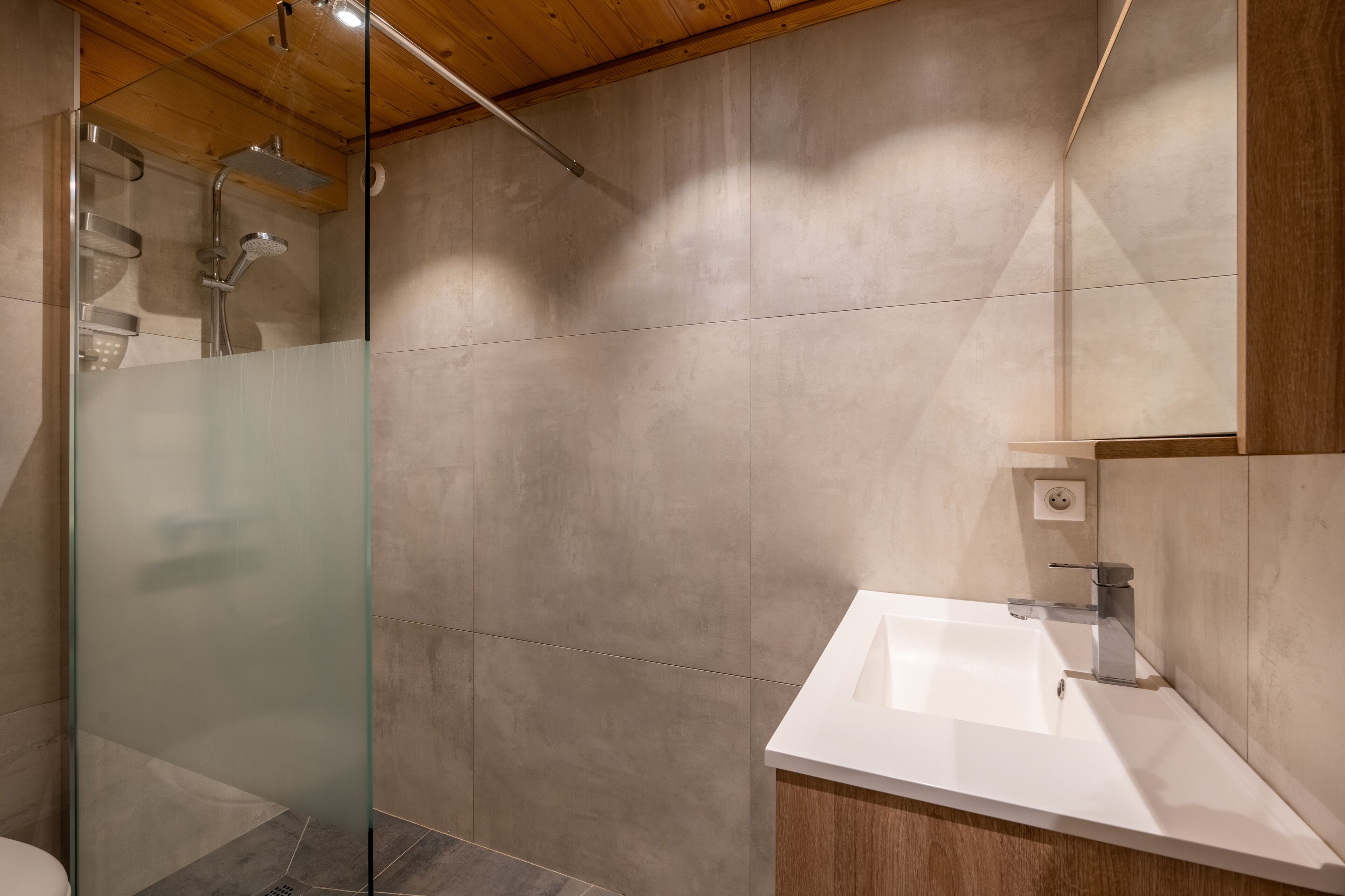 Une salle de bain moderne avec une douche à l’italienne en verre, un lavabo blanc et un meuble-vasque en bois.