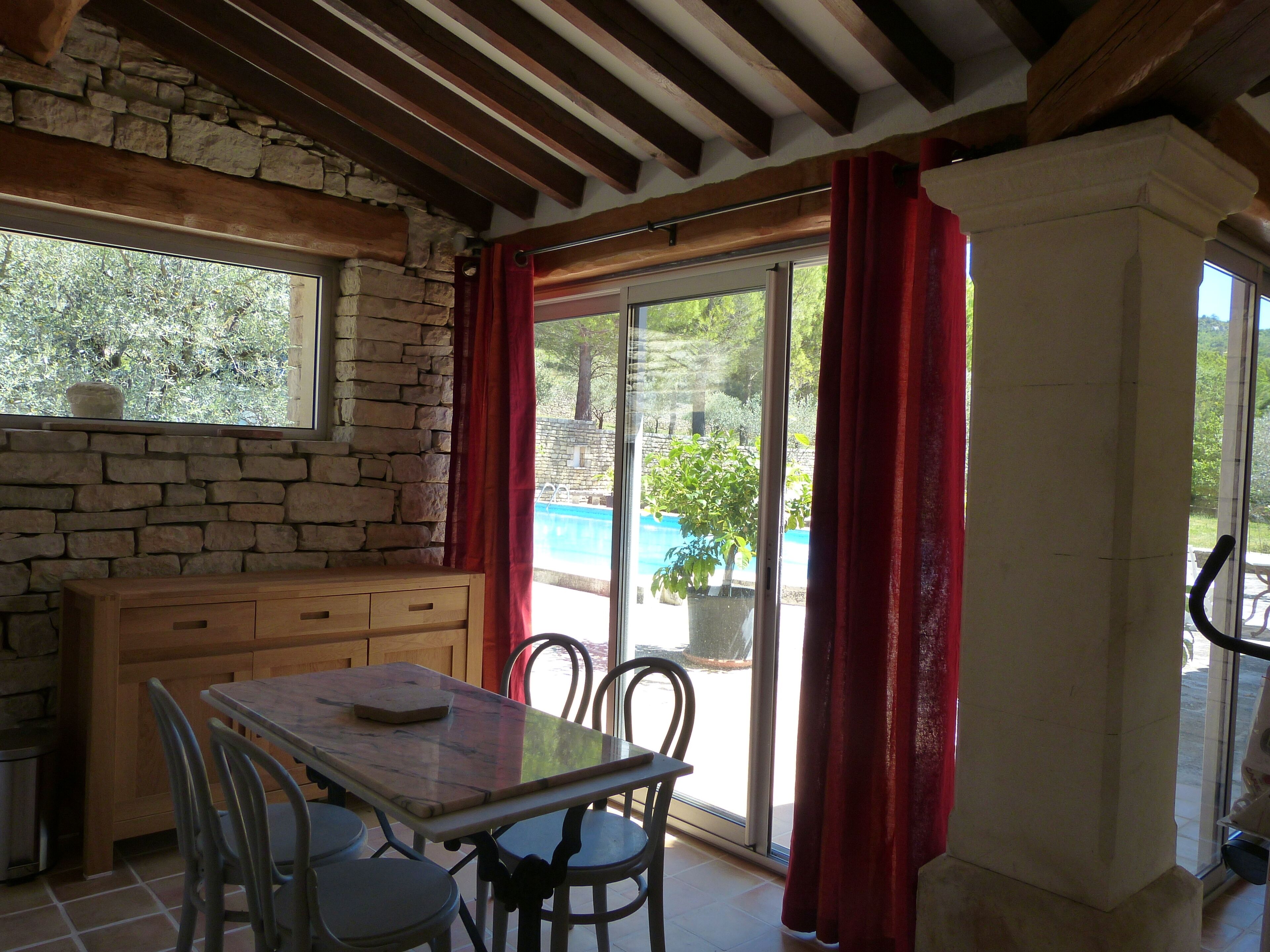 Un espace repas agrémenté d’un mur en pierre, de poutres en bois au plafond, d’une table avec des chaises et offrant une vue sur une piscine à travers de grandes portes vitrées.