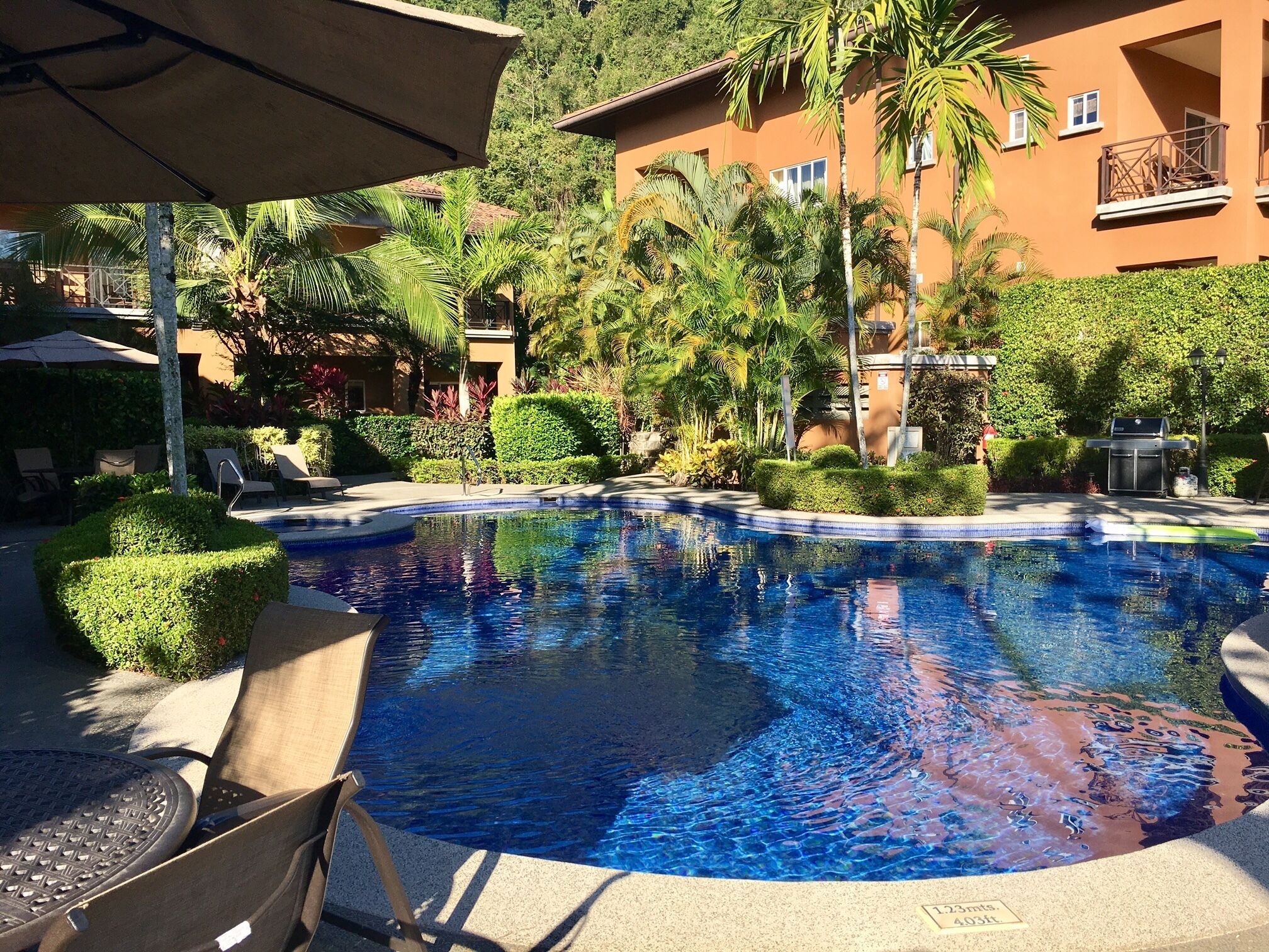 Una piscina con sillas de descanso y sombrillas, rodeada de exuberante vegetación y un edificio con balcones.