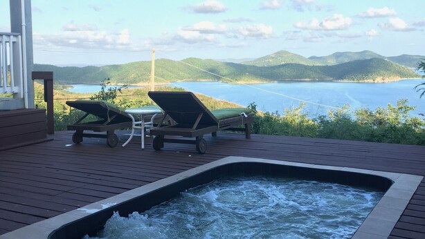 A wooden deck with a hot tub and lounge chairs overlooking a body of water and hills.