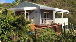 A two-story house with a balcony, surrounded by lush greenery and a hill in the background.