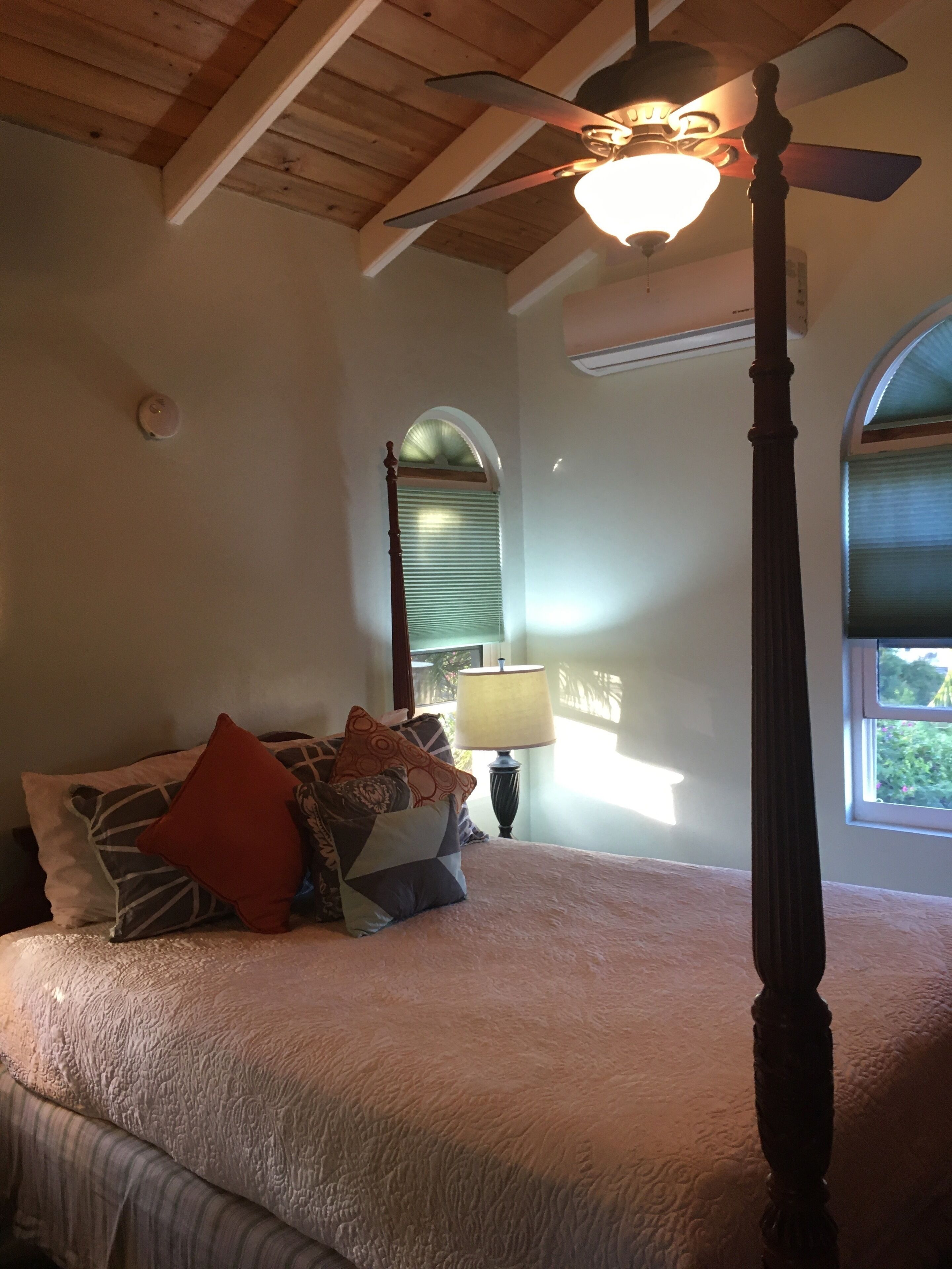 A bedroom with a four-poster bed, a ceiling fan, a window with blinds, and a lamp.