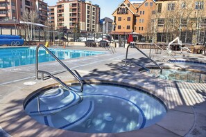 A swimming pool with a sunken center, surrounded by lounge chairs and umbrellas, with buildings in the background.