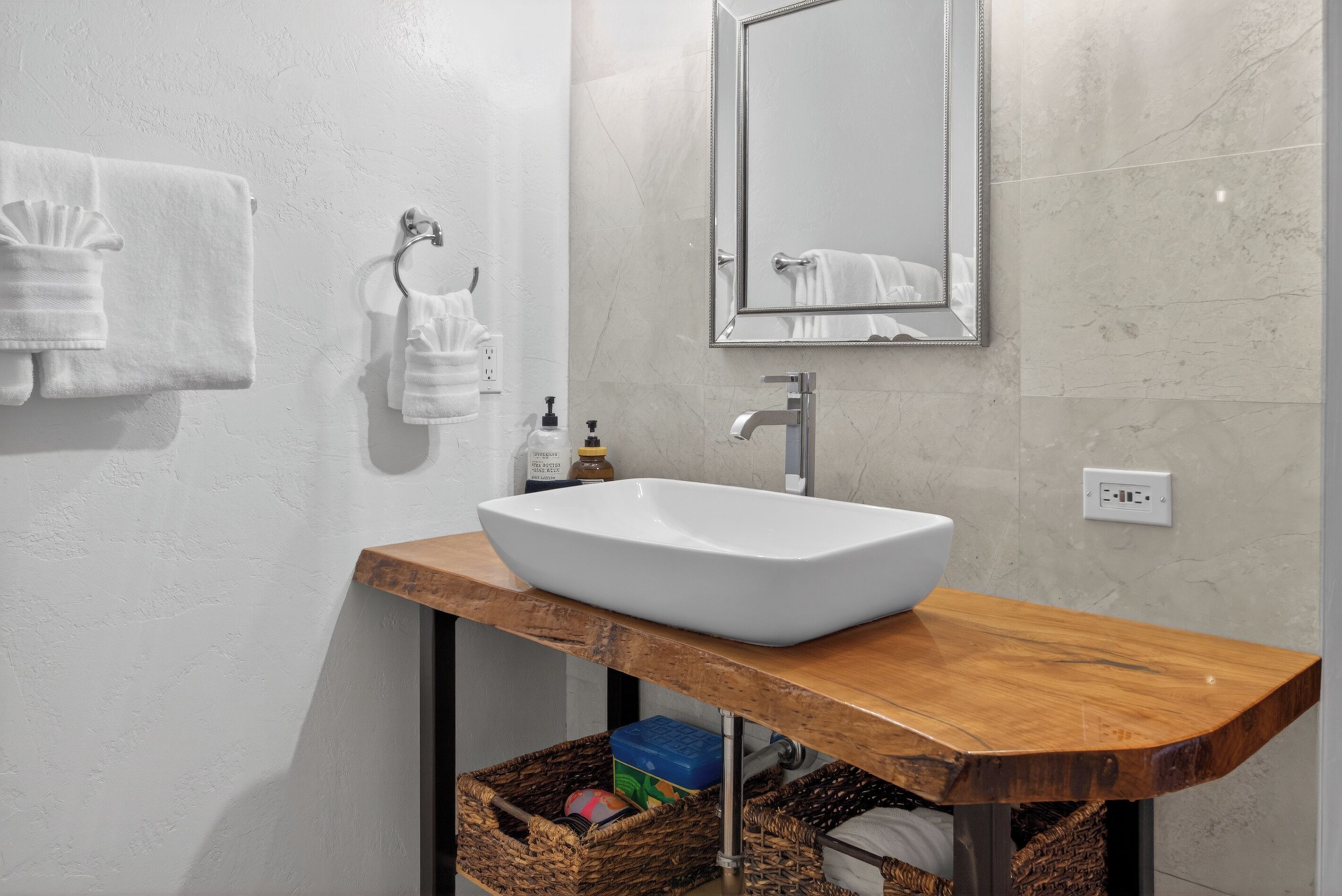 A bathroom with a white sink, wooden countertop, and a mirror above it. There are two towels hanging on hooks and a basket under the sink.