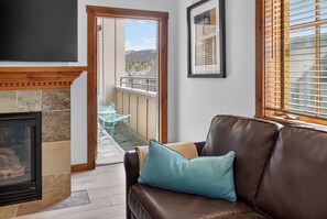 A living room with a fireplace, a sofa with cushions, a television, and a view of a balcony with outdoor furniture.