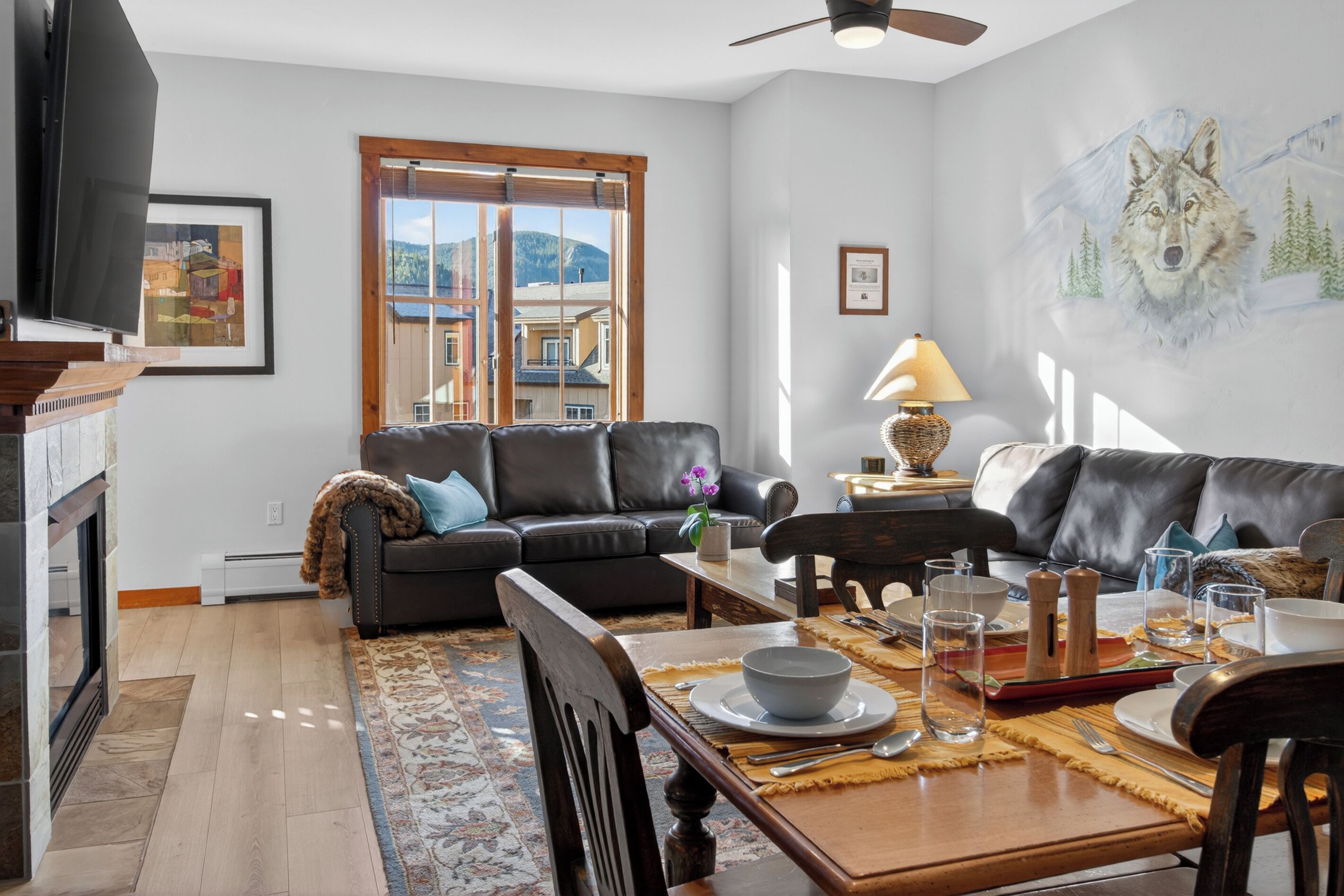 A modern living room with a fireplace, a dining area with a table set for two, and a large window with a view of buildings and mountains.