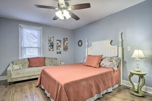 A bedroom with a bed, a ceiling fan, a window with blinds, a bedside table with a lamp, and a wall with framed pictures.