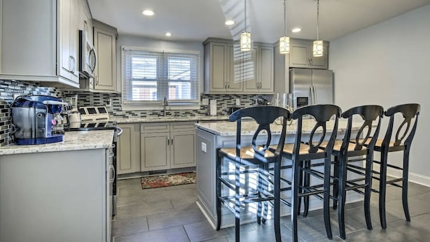 A modern kitchen with grey cabinets, a stainless steel refrigerator, a built-in island with bar stools, and pendant lighting. - Cottage on the Boulevard - Renovated, Historic Downtown Wendell-3 Beds, 2 Baths (Wendell)