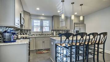 A modern kitchen with grey cabinets, a stainless steel refrigerator, a built-in island with bar stools, and pendant lighting.