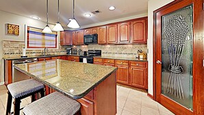 A kitchen with wooden cabinets, granite countertops, a central island with stools, a microwave, stove, and a decorative glass door. - Your Home Away From Home (Lexington)