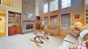 A spacious living room with a brick fireplace, a glass coffee table, and a beige sofa. There is a bookshelf filled with books and a television mounted on the wall. - Your Home Away From Home (Lexington)