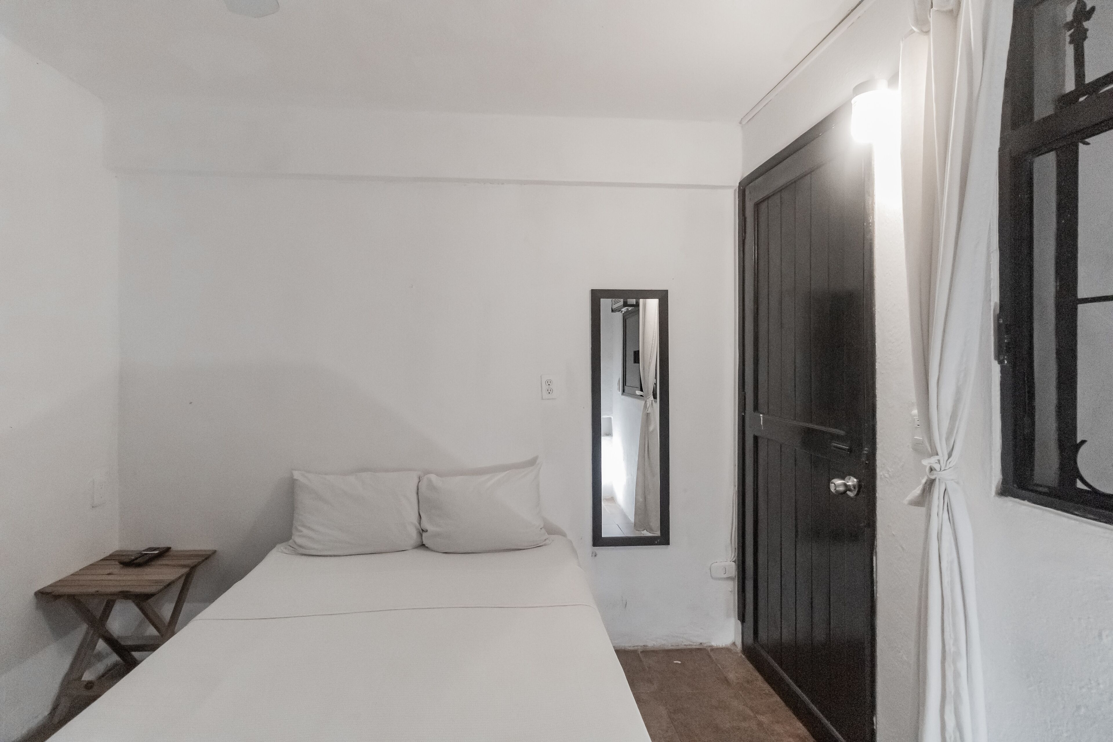 A small, simple room with a bed, a wooden nightstand, a mirror, and a dark door.