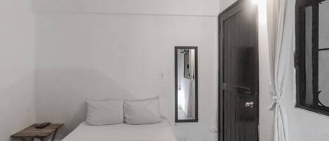 A small, simple room with a bed, a wooden nightstand, a mirror, and a dark door.