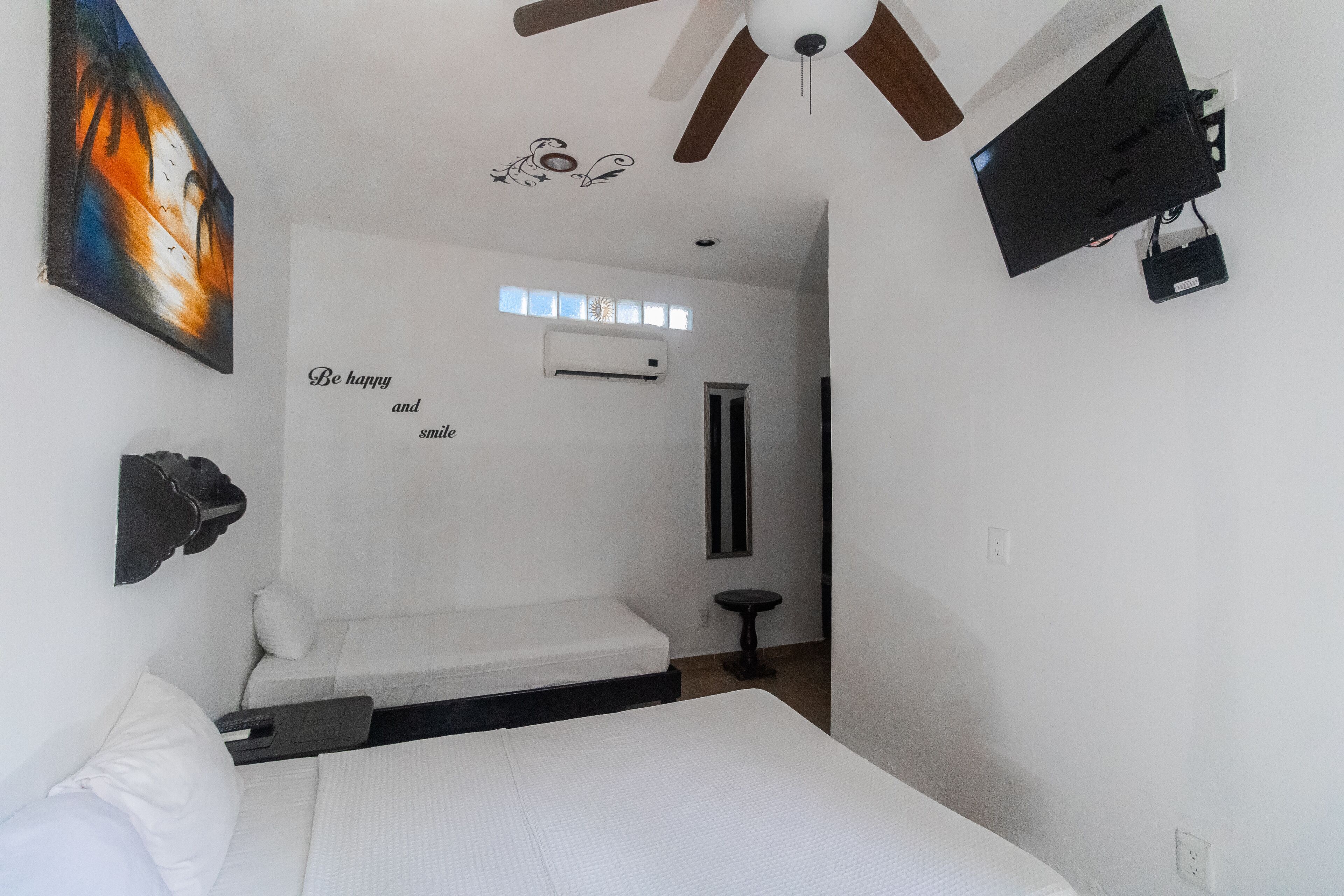A small, white room with a bed, a ceiling fan, an air conditioning unit, and a wall-mounted TV.