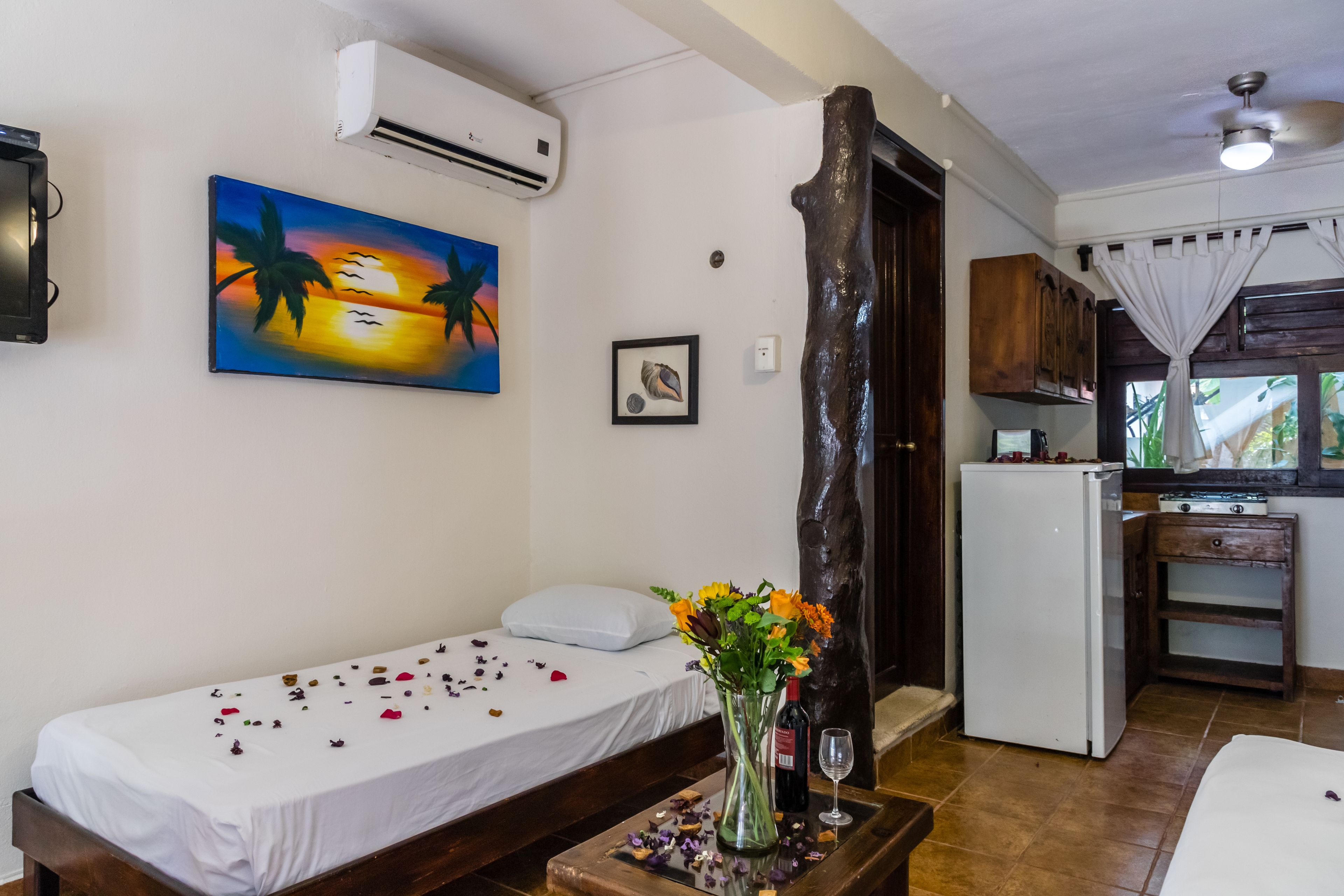 Una habitación con una cama, una mesita con un florero y una copa de vino, un cuadro de un atardecer y una zona de cocina con un refrigerador y armarios.