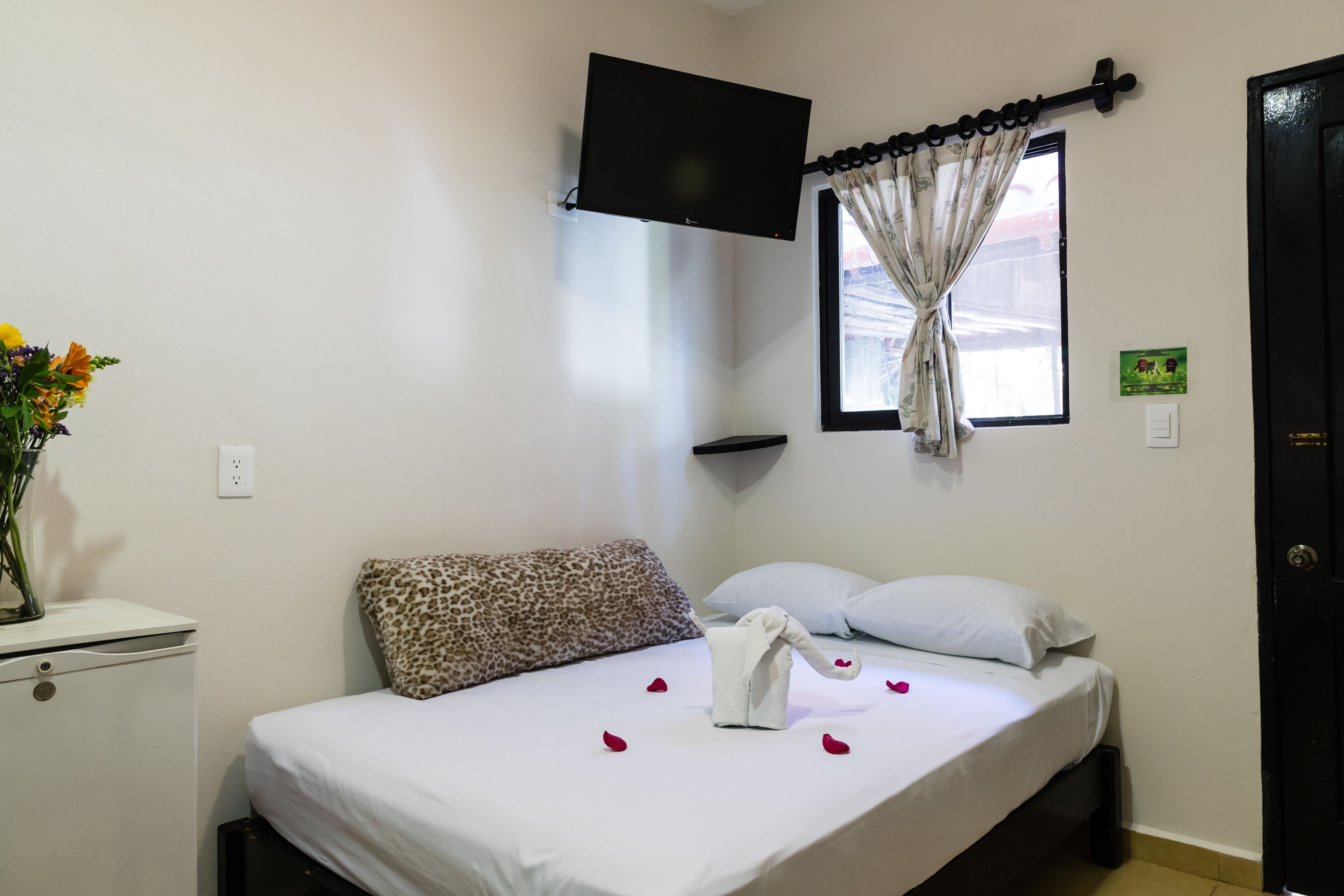 A hotel room with a bed, a TV mounted on the wall, a window with curtains, and a small desk with a vase of flowers.