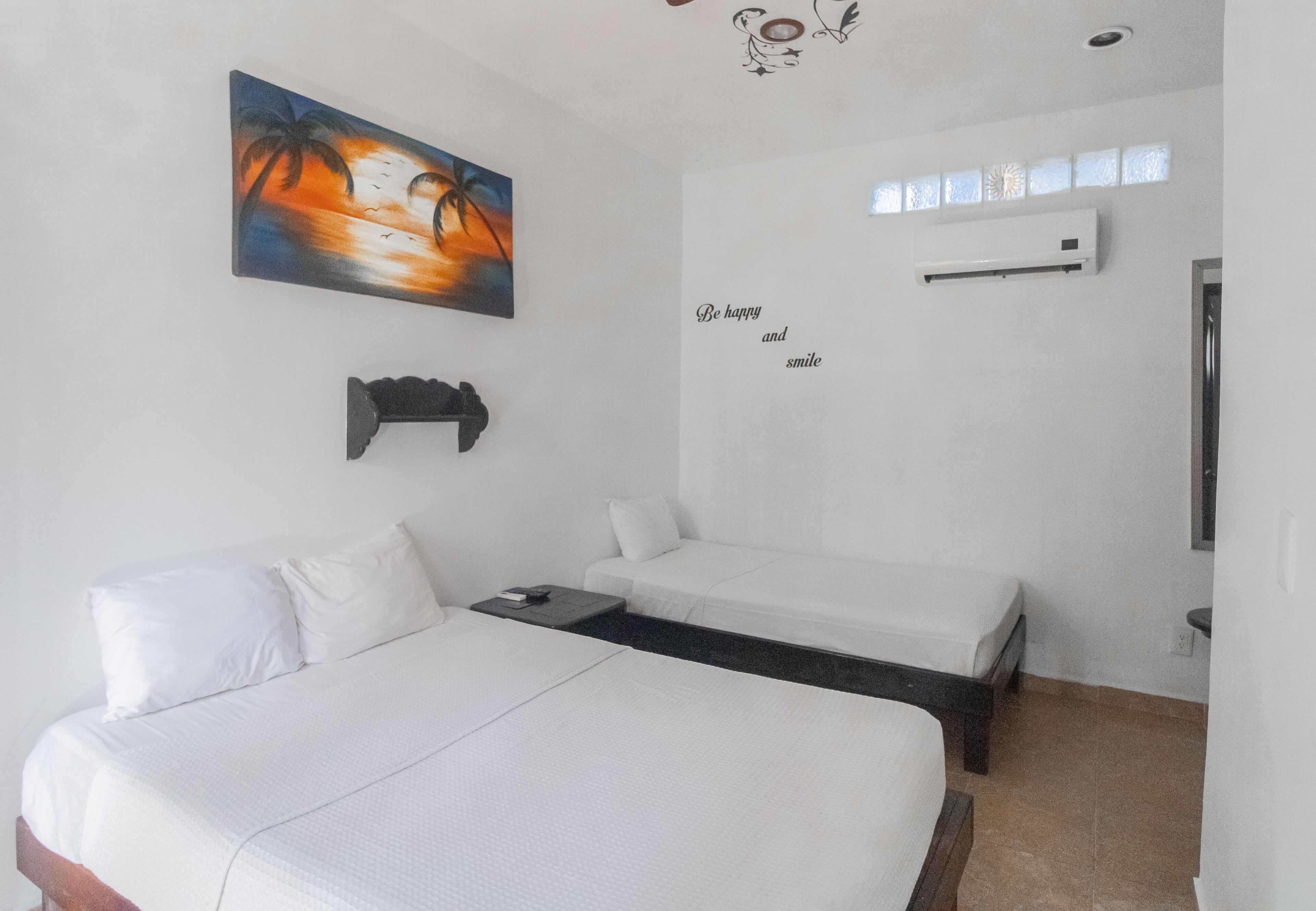 A hotel room with two single beds, a painting of a sunset, a motivational quote on the wall, and an air conditioning unit.
