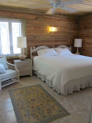 A wooden room with a bed, a chair, a nightstand, a lamp, and a window.