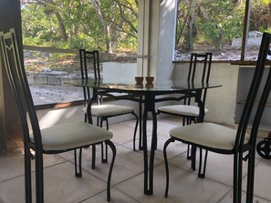 A glass-top table with four chairs, a small table with two decorative items, and a view of trees and greenery outside.