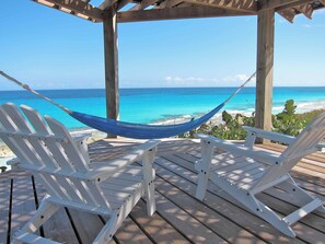 Eine Holzterrasse mit weißen Liegestühlen und einer Hängematte mit Blick auf einen Strand und klares, blaues Wasser.