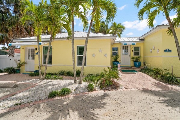 A yellow building with a white door and windows, surrounded by palm trees and a gravel driveway. -  Cozy Cottage, 1 minute walk to Beach (Clearwater)