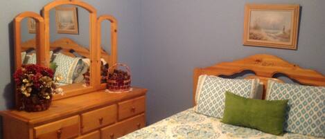 A bedroom with a wooden dresser, a bed with patterned bedding, a green pillow, and a framed picture on the wall.