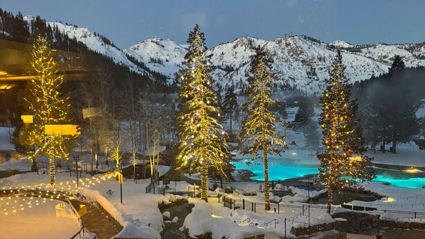 Un espacio exterior cubierto de nieve, con árboles de Navidad e iluminación, una piscina con agua azul y montañas al fondo.