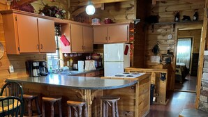 Cocina de estilo rústico tipo cabaña, con gabinetes de madera, una isla central con taburetes, un refrigerador y un letrero que dice 'Welcome'.
