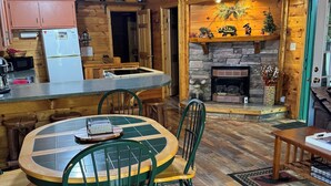 Un acogedor interior de cabaña con una mesa y sillas de madera, chimenea de piedra y una zona de cocina con refrigerador y microondas.