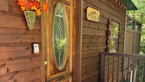 Entrada a una cabaña de madera con puerta de vidrio decorativa, felpudo y un letrero que dice 'Bear About'.