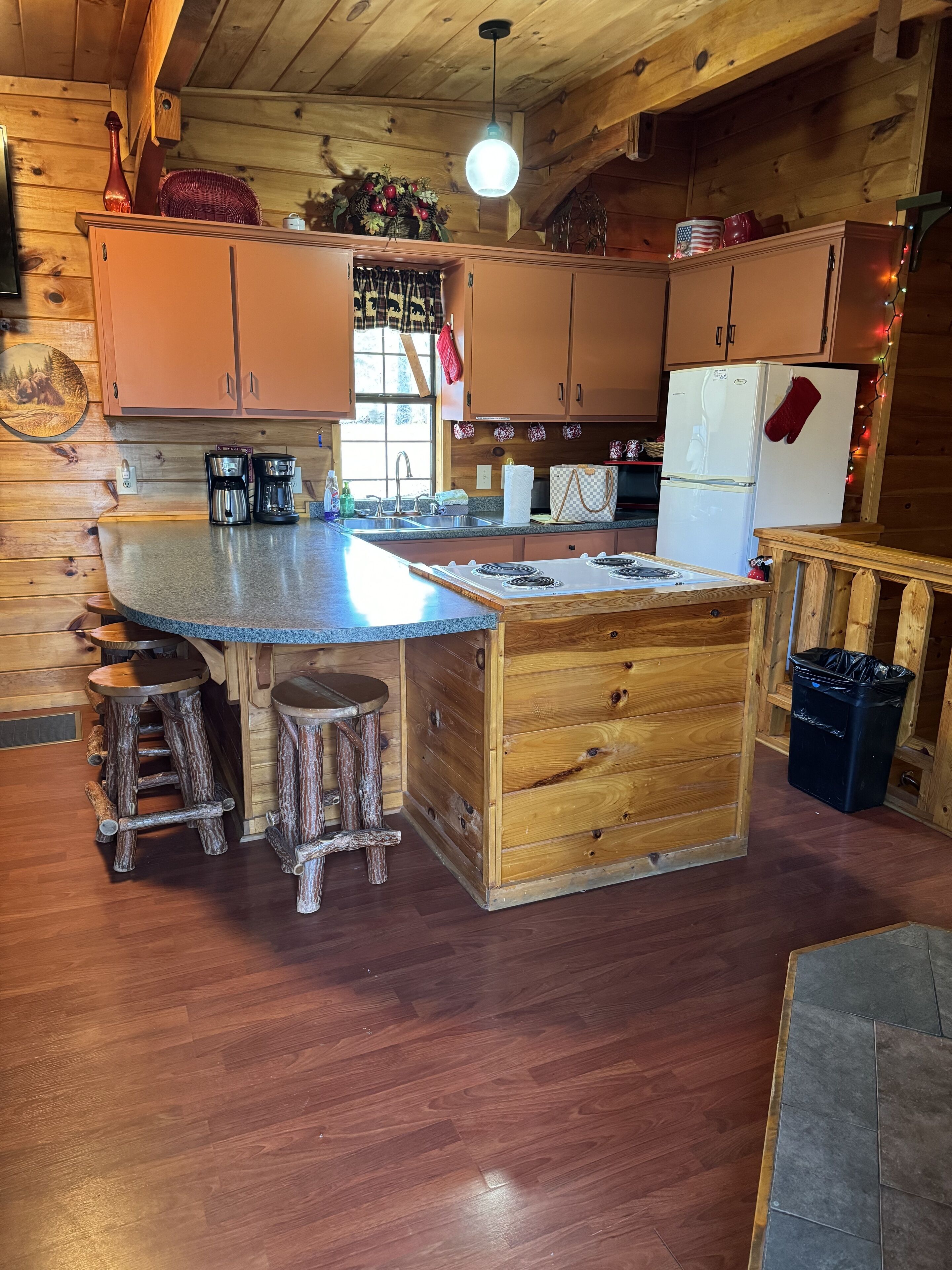 A rustic kitchen with wooden cabinets, a central island with stools, a refrigerator, and a sink.