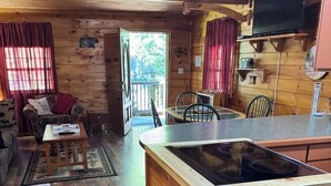 Interior de una cabaña de madera con cocina, mesa de comedor y sala de estar con sofá y televisor.