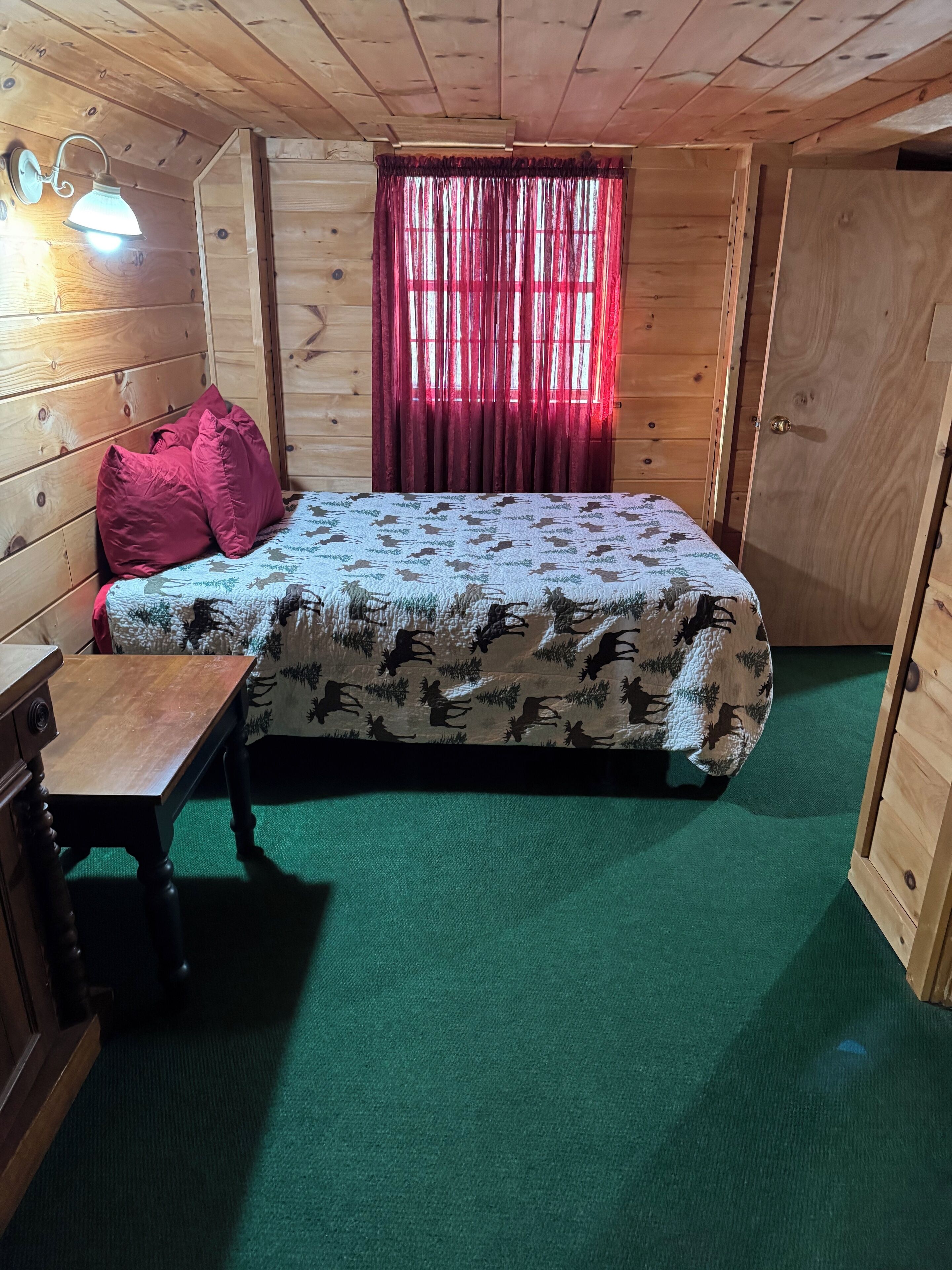 A wooden cabin room with a bed, a small table, a lamp, and a window with curtains.