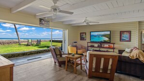 A living room with a large TV, wooden furniture, and a view of the ocean. - Oceanfront Hawaiian-style Beach House (Waimea)