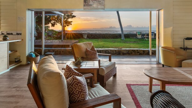 A cozy living room with a view of the sunset over the ocean, furnished with a sofa, chairs, and a coffee table. - Oceanfront Hawaiian-style Beach House (Waimea)