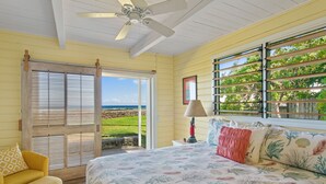 A bedroom with a bed, a yellow armchair, a ceiling fan, and a view of the ocean through a sliding door. - Oceanfront Hawaiian-style Beach House (Waimea)