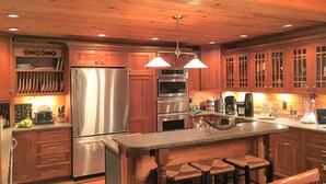 A kitchen with wooden cabinets, a stainless steel refrigerator, an oven, and a coffee maker. There are three bar stools and a wooden ceiling. - Fabulous Log Cabin: the perfect getaway with new  pickleball court and pool! (Monterey)