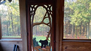 A wooden cabin with a large wooden door featuring a bear illustration, a wicker chair, and a view of a forest. - Fabulous Log Cabin: the perfect getaway with new  pickleball court and pool! (Monterey)