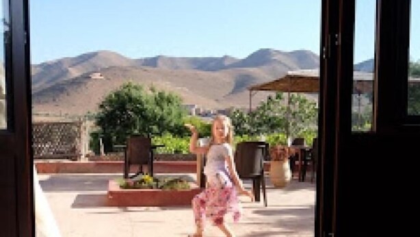 Un enfant vêtu d’une robe blanche à motifs floraux traverse un seuil de porte ouvert.