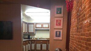 A narrow room with a wooden floor, a table with four stools, a brick wall, and a kitchen visible through an open doorway.