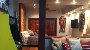A room with a wooden floor, a grey sofa with red and white pillows, a brick wall, a wooden door, and a ceiling fan.