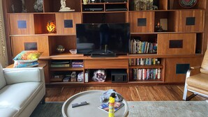Uma sala de estar com uma televisão de tela grande, estantes de madeira com objetos decorativos, livros e uma mesa de centro com controles remotos.
