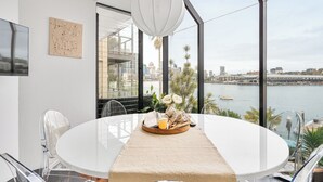 Un moderno comedor con una mesa redonda, sillas transparentes, un mantel blanco, una lámpara colgante y vistas a un paisaje urbano y al agua.