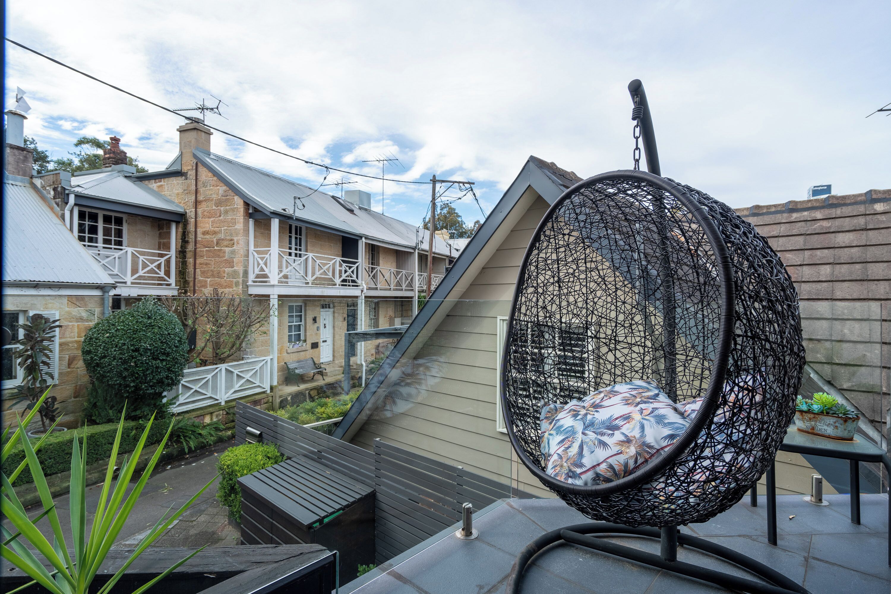 A hanging chair with a patterned cushion, a stone house with a white balcony, and a small table with a potted plant.