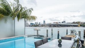 Piscina en la azotea con barandilla de vidrio, mobiliario de mimbre y vistas al puerto deportivo y al horizonte de la ciudad.